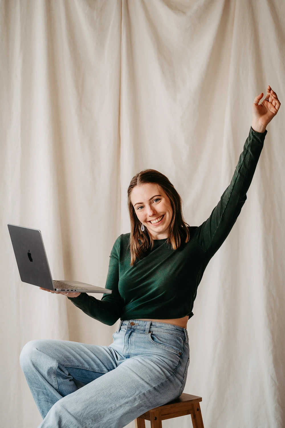 Anna Proprentner auf einem Hocker mit ihrem Laptop, sie wirft die Hände in die Höhe. Sie wirkt fröhlich und motiviert und voller Elan.
