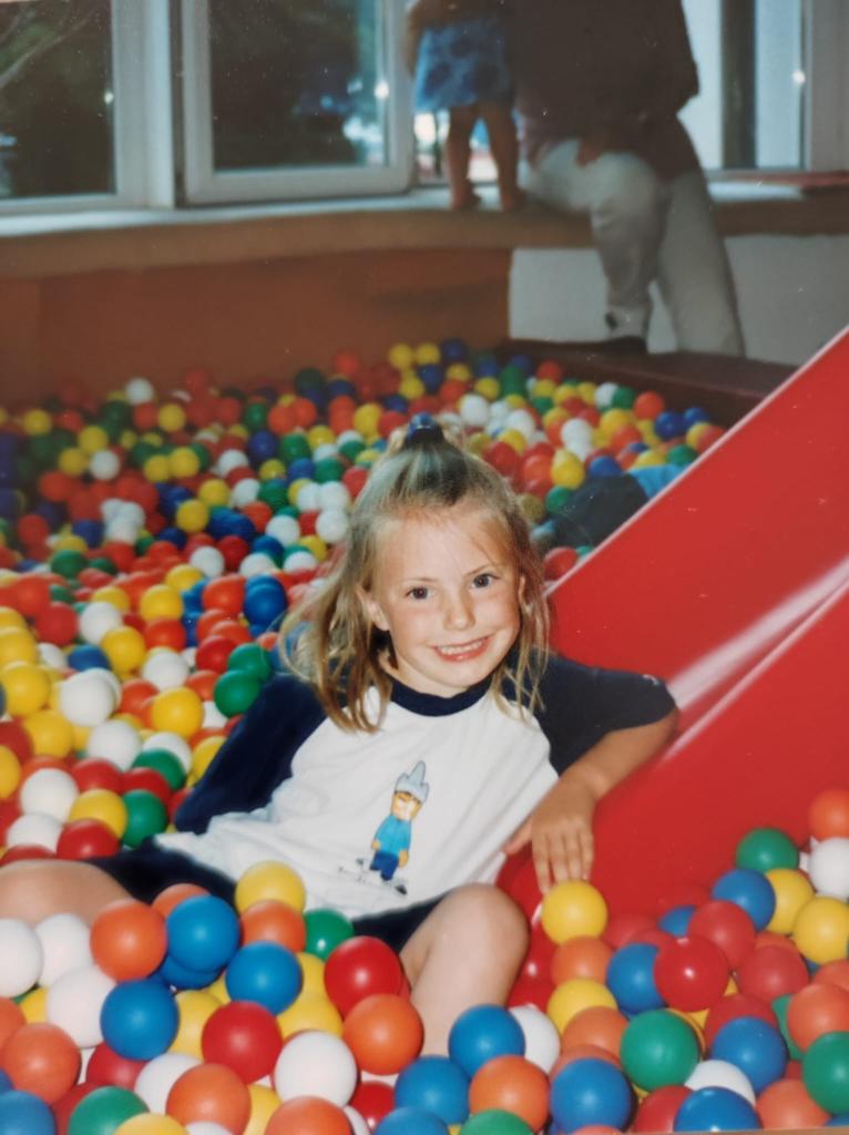 Anna als kleines Mädchen. Sie lächelt keck. Sie ist gerade über eine Rutsche in ein Bällebad gerutscht.